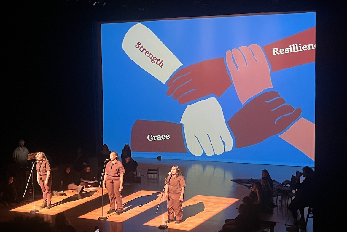 Three participants in dark clothing on stage at the Glorya Kaufman Theater, with a projection of linked hands on a screen behind them