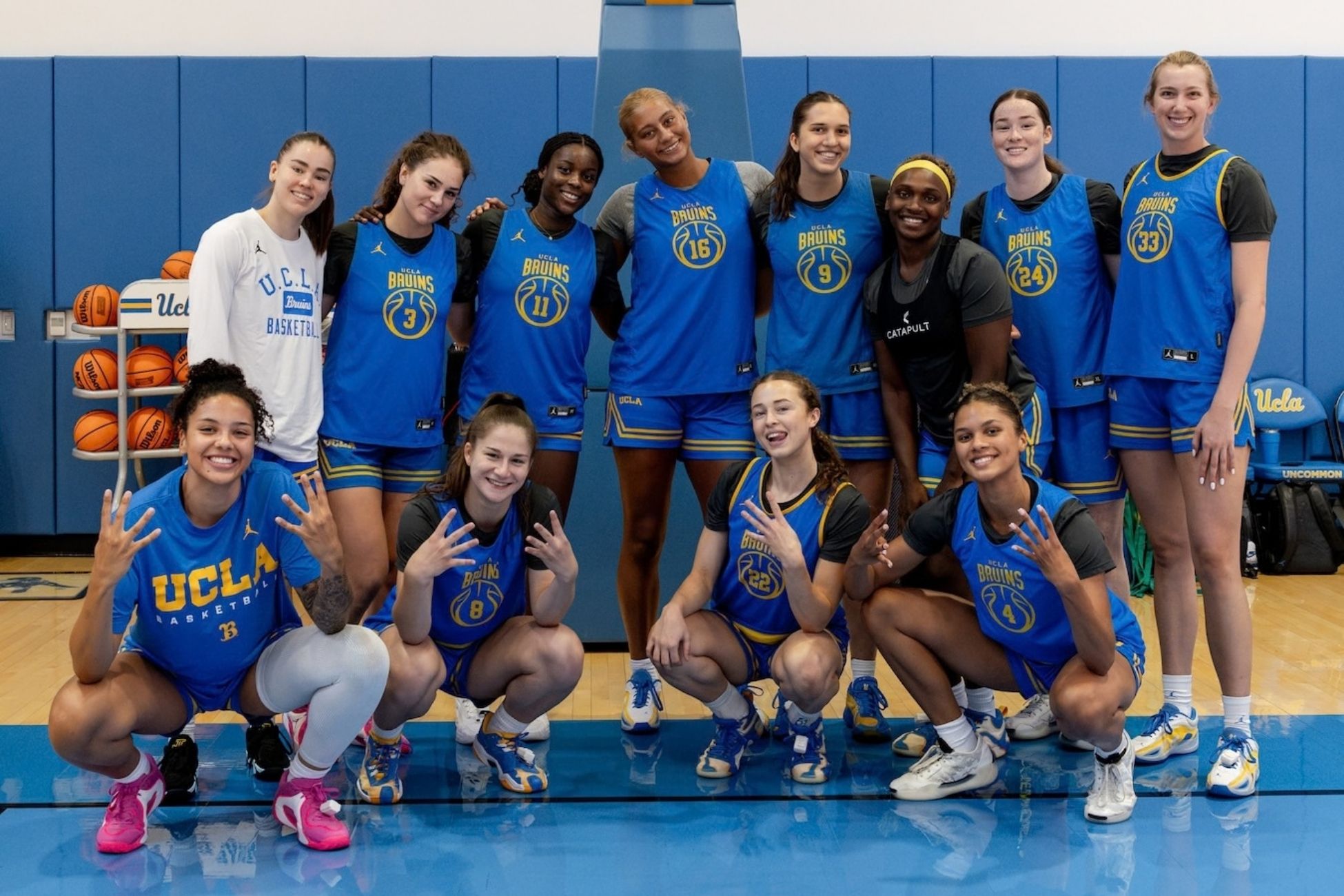 A group of UCLA women’s basketball players pose together in blue and gold Bruins uniforms inside a gym, smiling and making hand gestures, with basketballs and blue padded walls in the background.