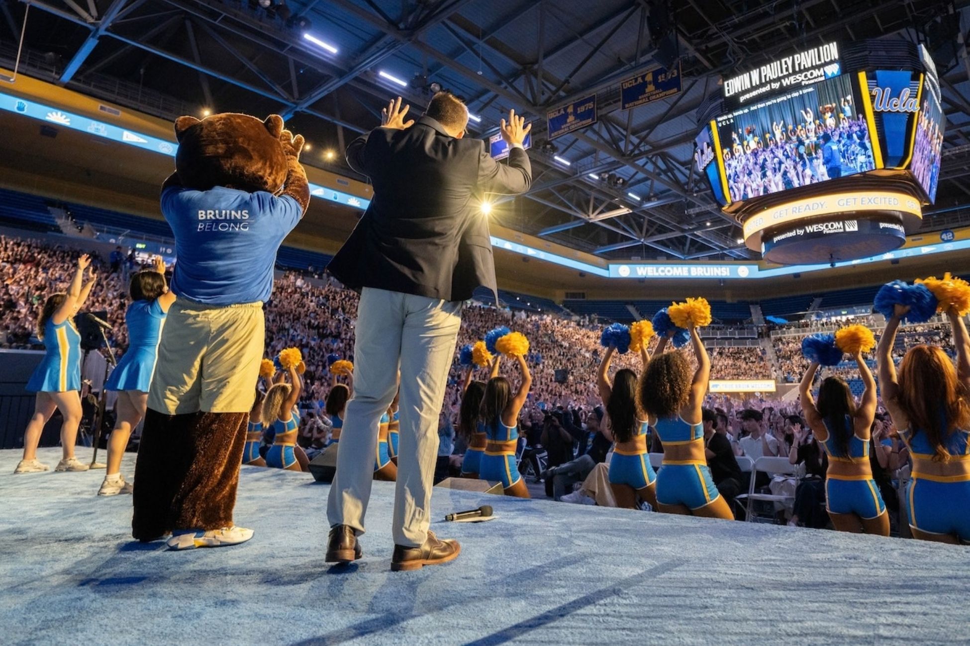 A UCLA mascot and a man lead a cheering crowd at Pauley Pavilion, surrounded by UCLA spirit squad members waving blue and gold pom-poms.