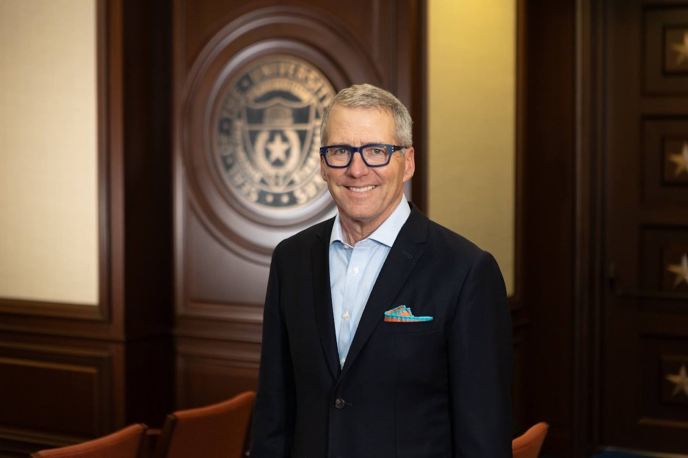 A man wearing glasses and a suit stands smiling in front of a polished wooden panel backdrop.