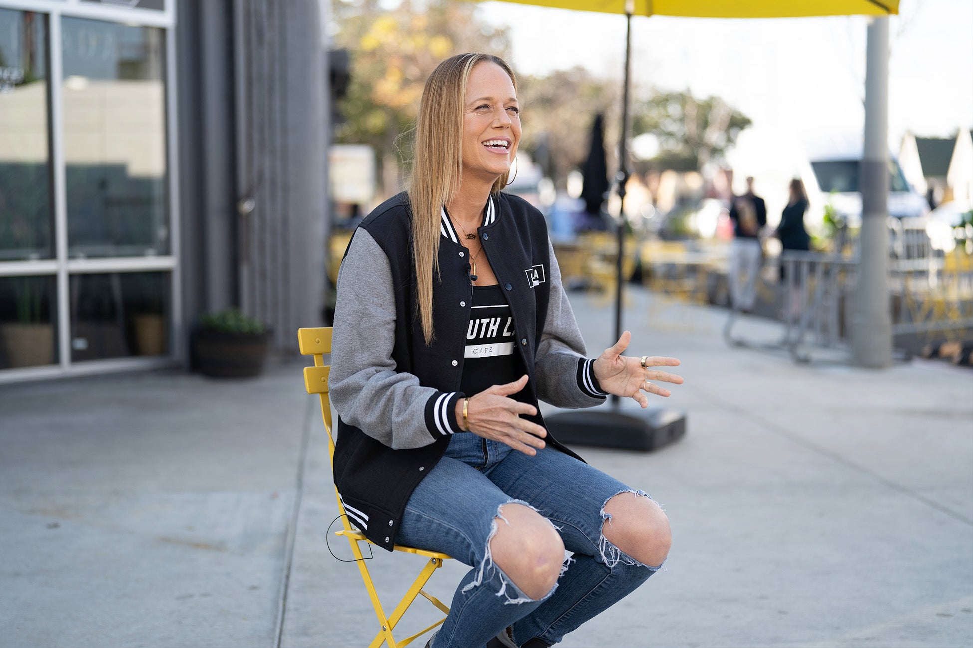 A woman sits outdoors on a yellow chair, smiling and gesturing as she speaks with a casual and animated demeanor.