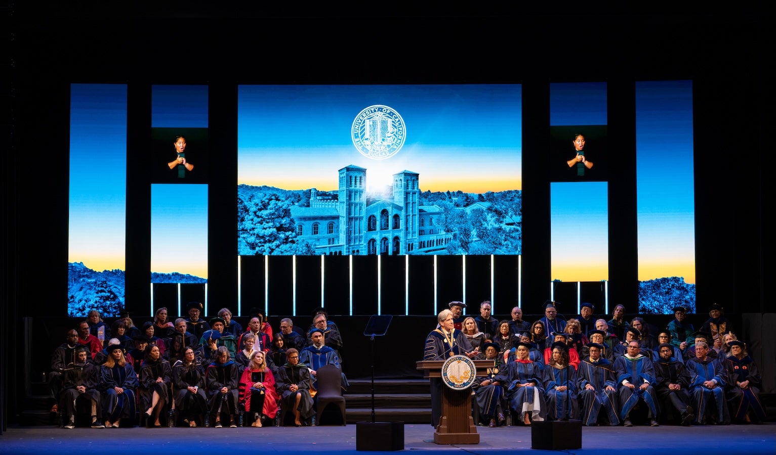 A large group of people sit on stage, with a large screen displaying visuals behind them and Chancellor Frenk speaks at a podium.