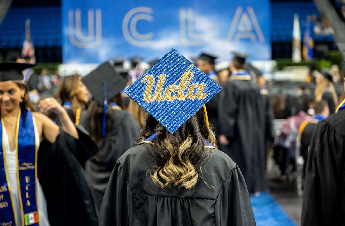 A graduate in a gown and cap with UCLA decorated on it stands facing a bustling crowd of graduates