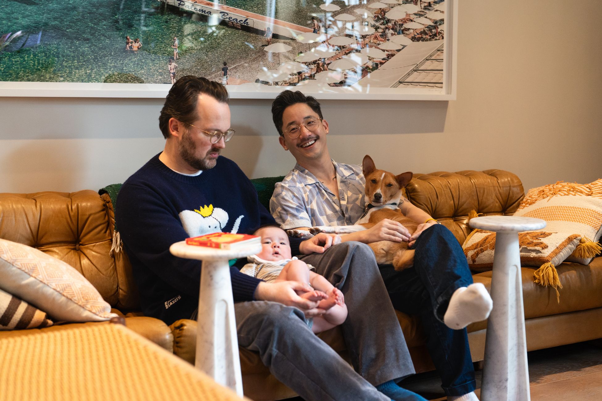 Two men sit together on a tan leather couch, one holding a baby and the other holding a dog, all appearing relaxed and content in a cozy home setting. A framed beach photograph hangs on the wall behind them, and children&#039;s books rest on small white tables in front.