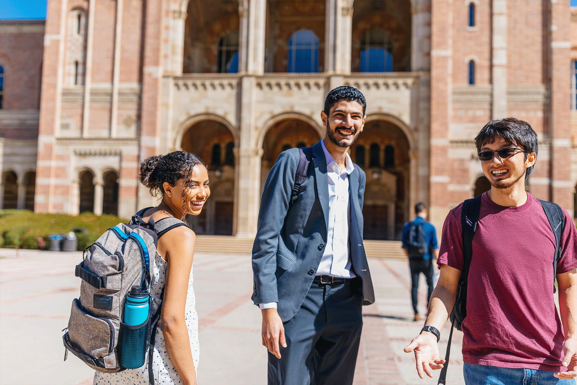 Three students, one with backpack, stand in front of Royce Hall and smile to camera