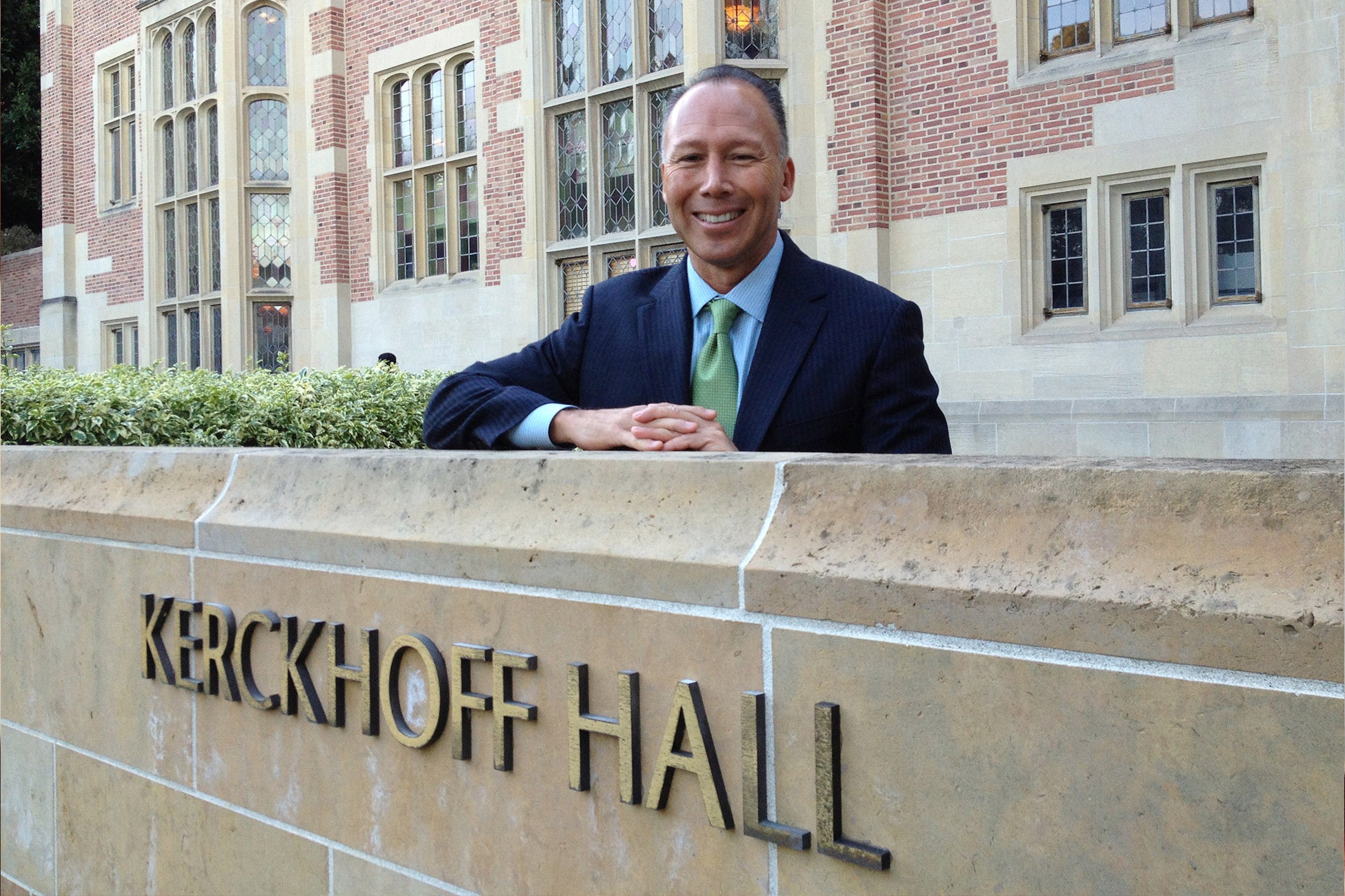 A smiling man in a suit and tie stands behind a stone wall with the words &quot;KERCKHOFF HALL&quot; engraved on it.