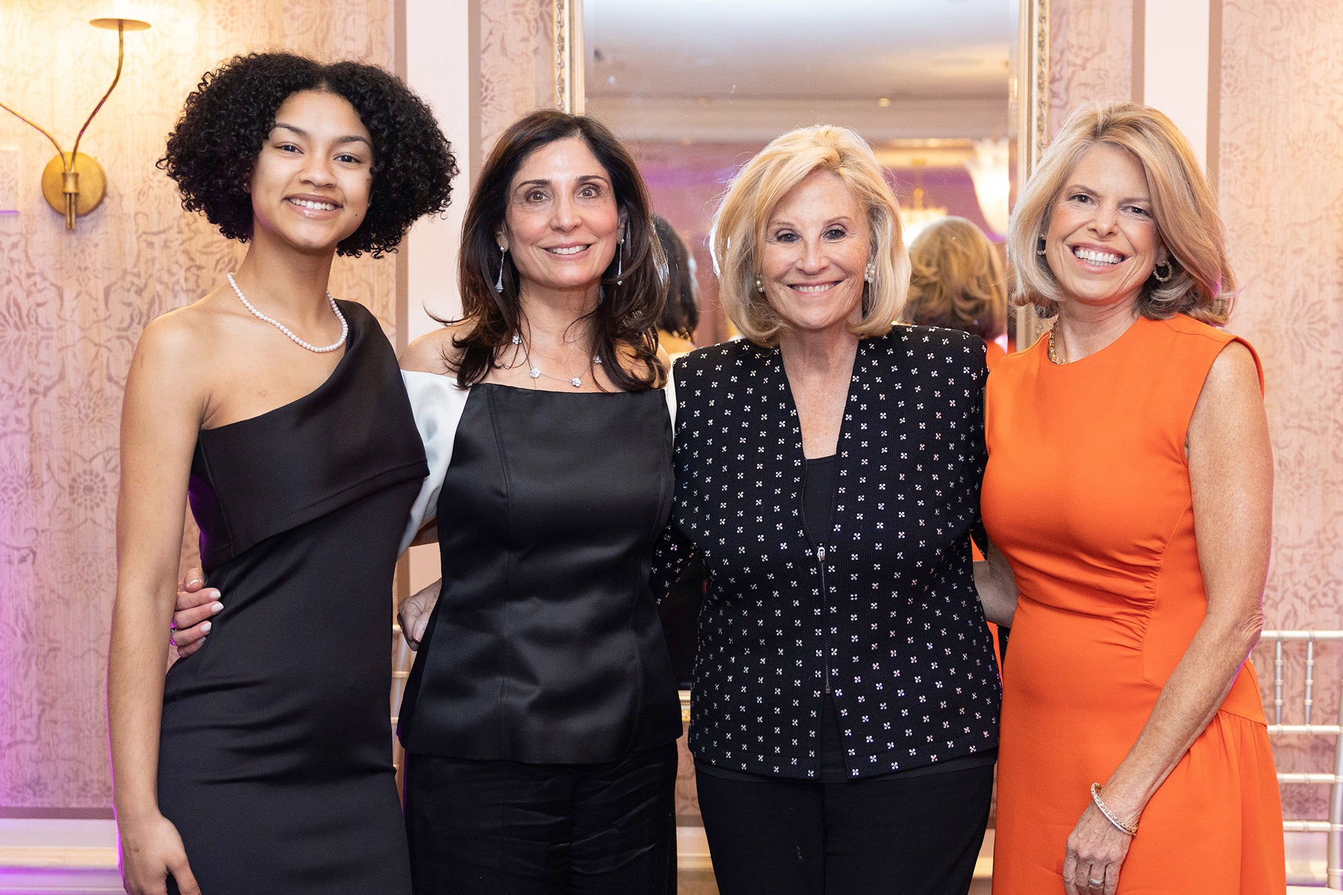 Four women pose together and smile at a formal event, standing in front of a mirrored wall with warm lighting. They are dressed in elegant evening attire, suggesting a celebratory or gala occasion.
