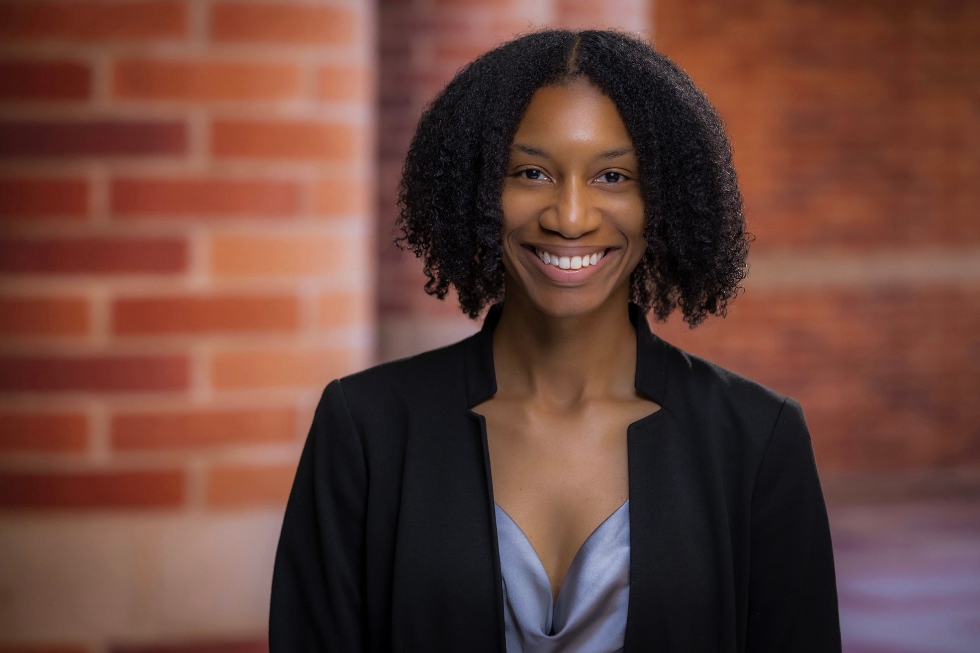 Woman with chin-length dark coils, black blazer stands in front of brick building and smiles