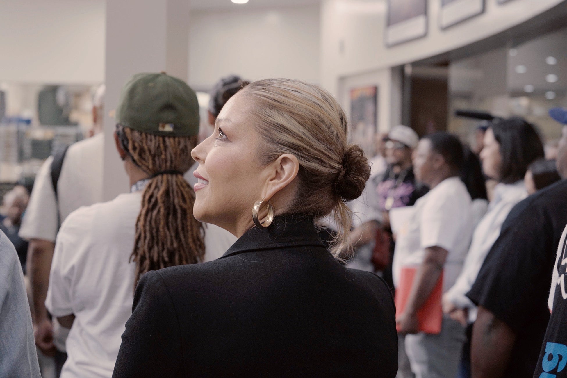 Woman in a black blazer and gold hoop earrings looks upward while standing among a group of people indoors.