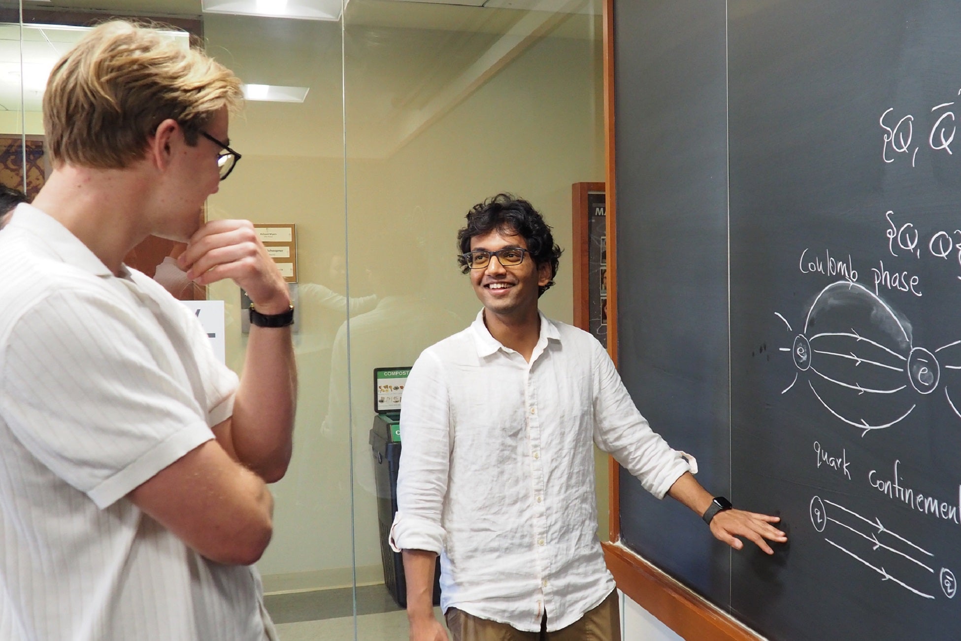 Two men discuss a diagram and equation on a blackboard.