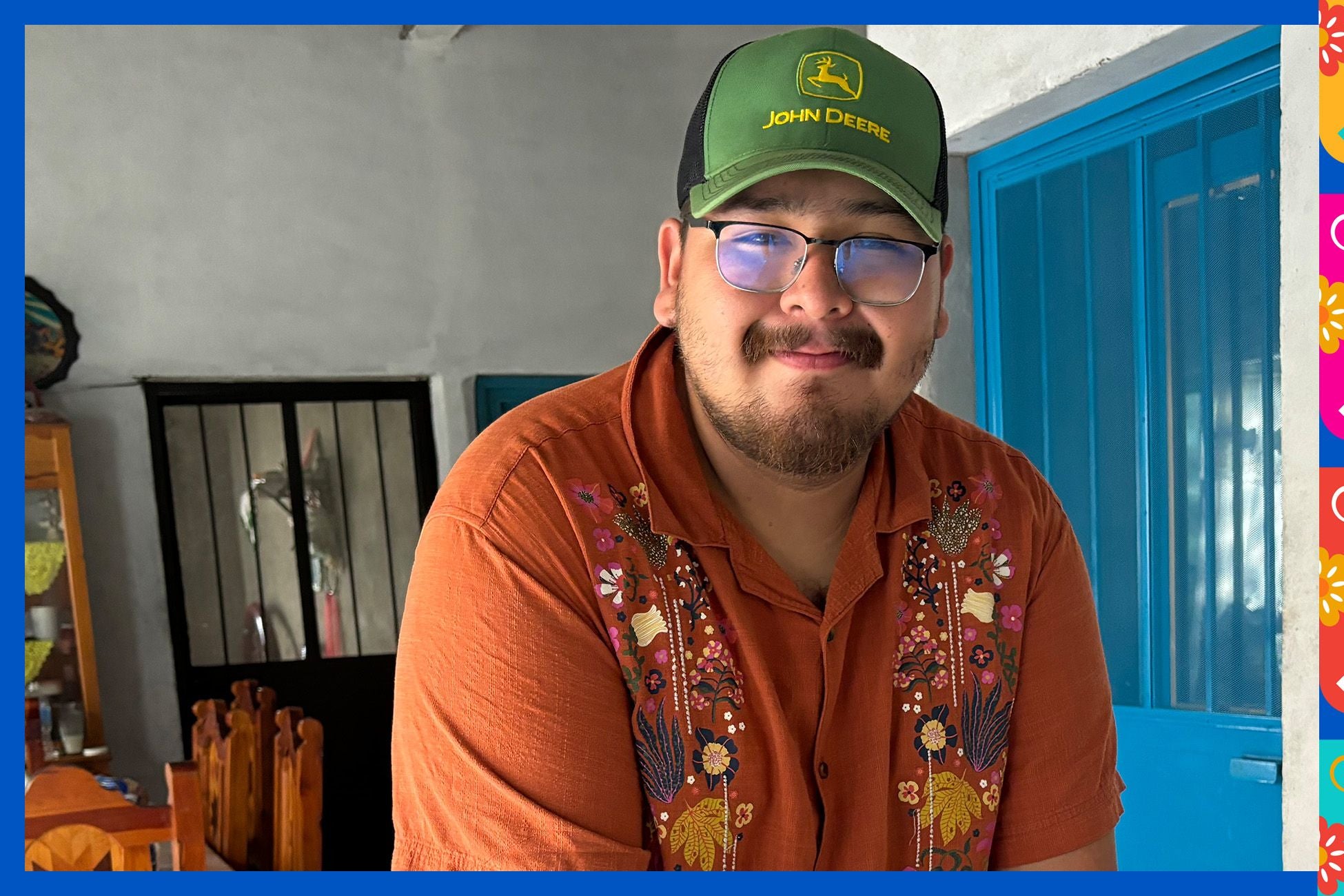 A man wearing glasses and a hat smiles against a bright, multicolored wall.