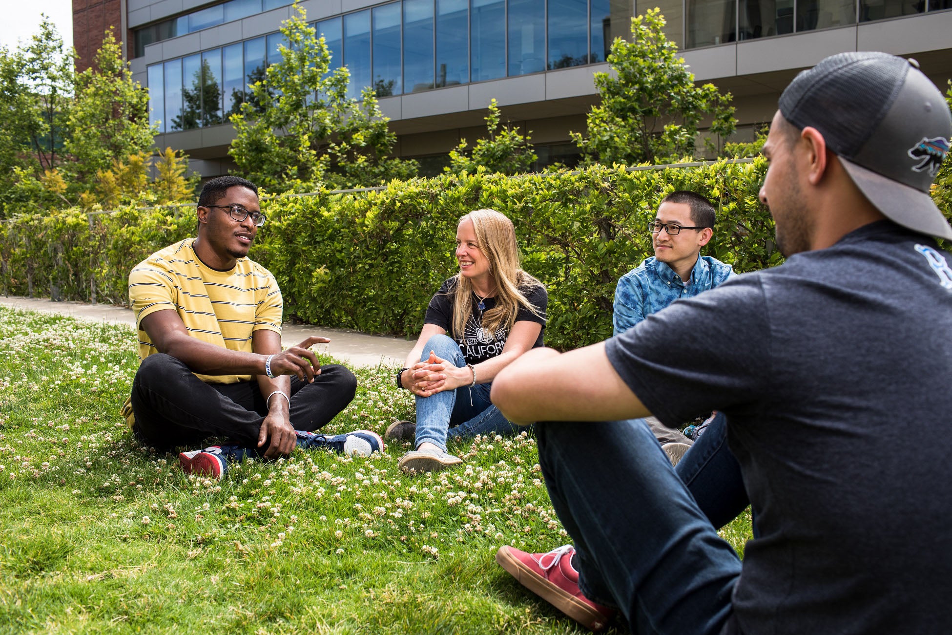 Four people, one on right with back to camera, sit on grass facing each other and talk