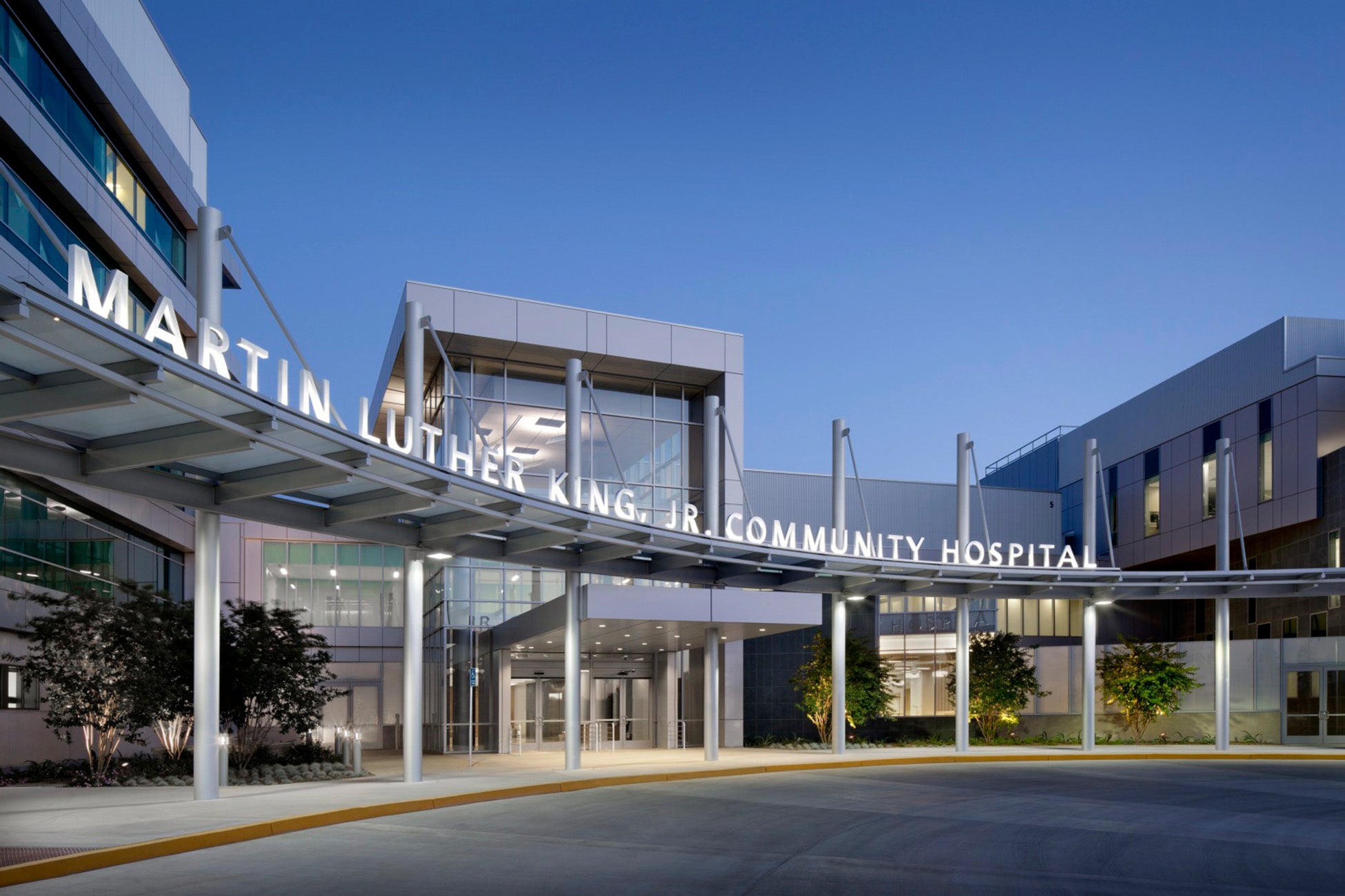 Exterior view of the modern entrance to Martin Luther King, Jr. Community Hospital at dusk, featuring large glass windows, metal beams, and illuminated signage above a curved driveway.