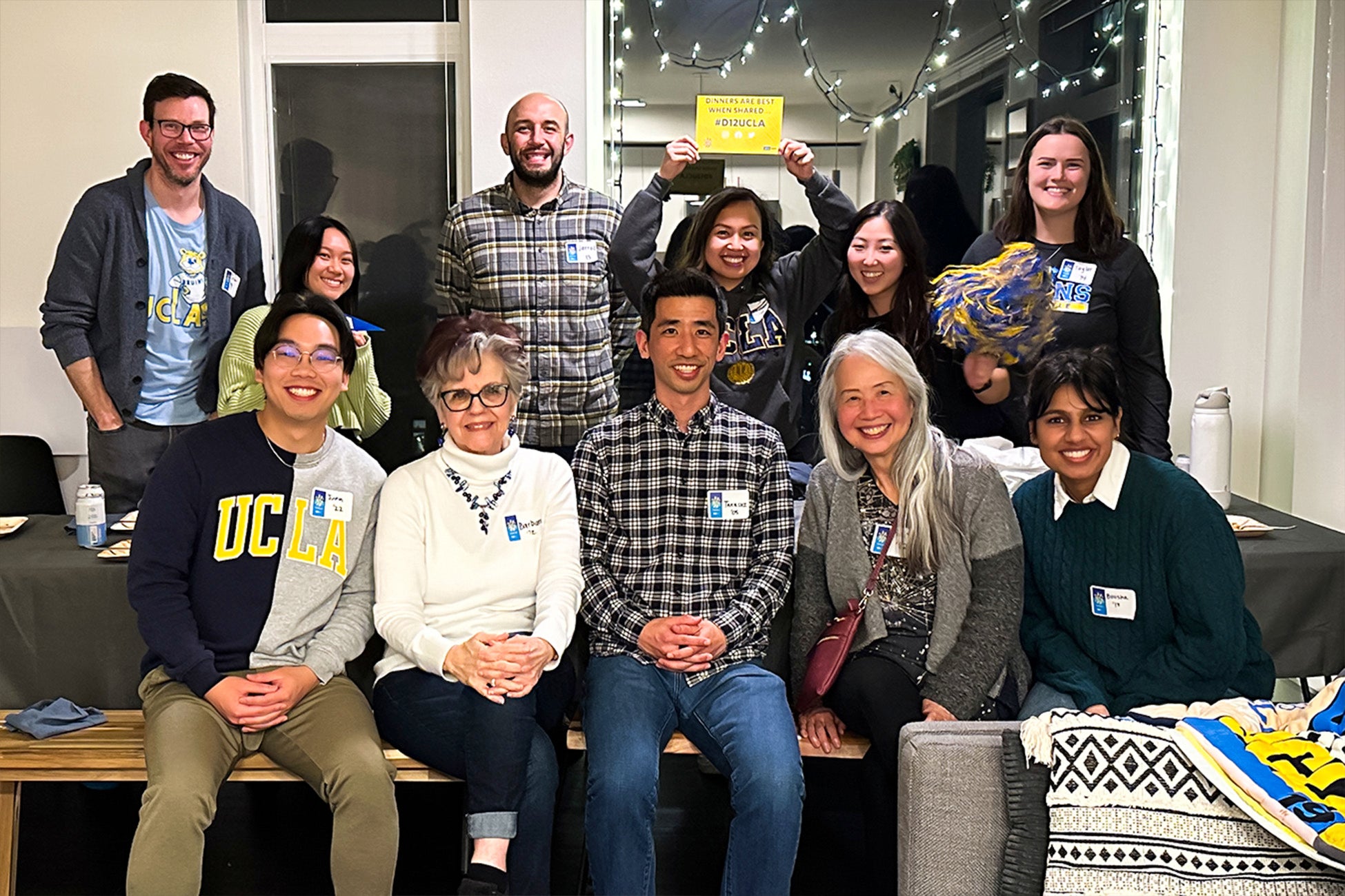 A group of UCLA alumni and community members pose indoors, smiling and wearing UCLA apparel at a casual gathering