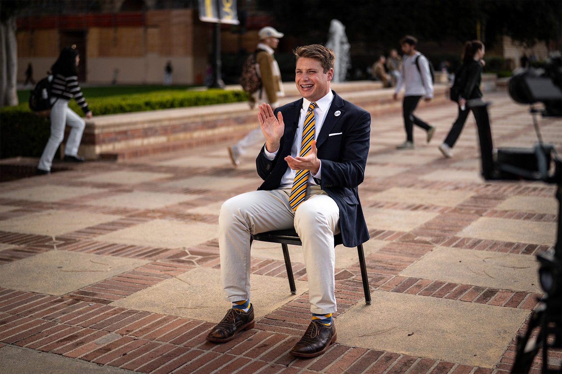 A person in business attire sits on a chair outdoors on campus, smiling and clapping as students walk by in the background.