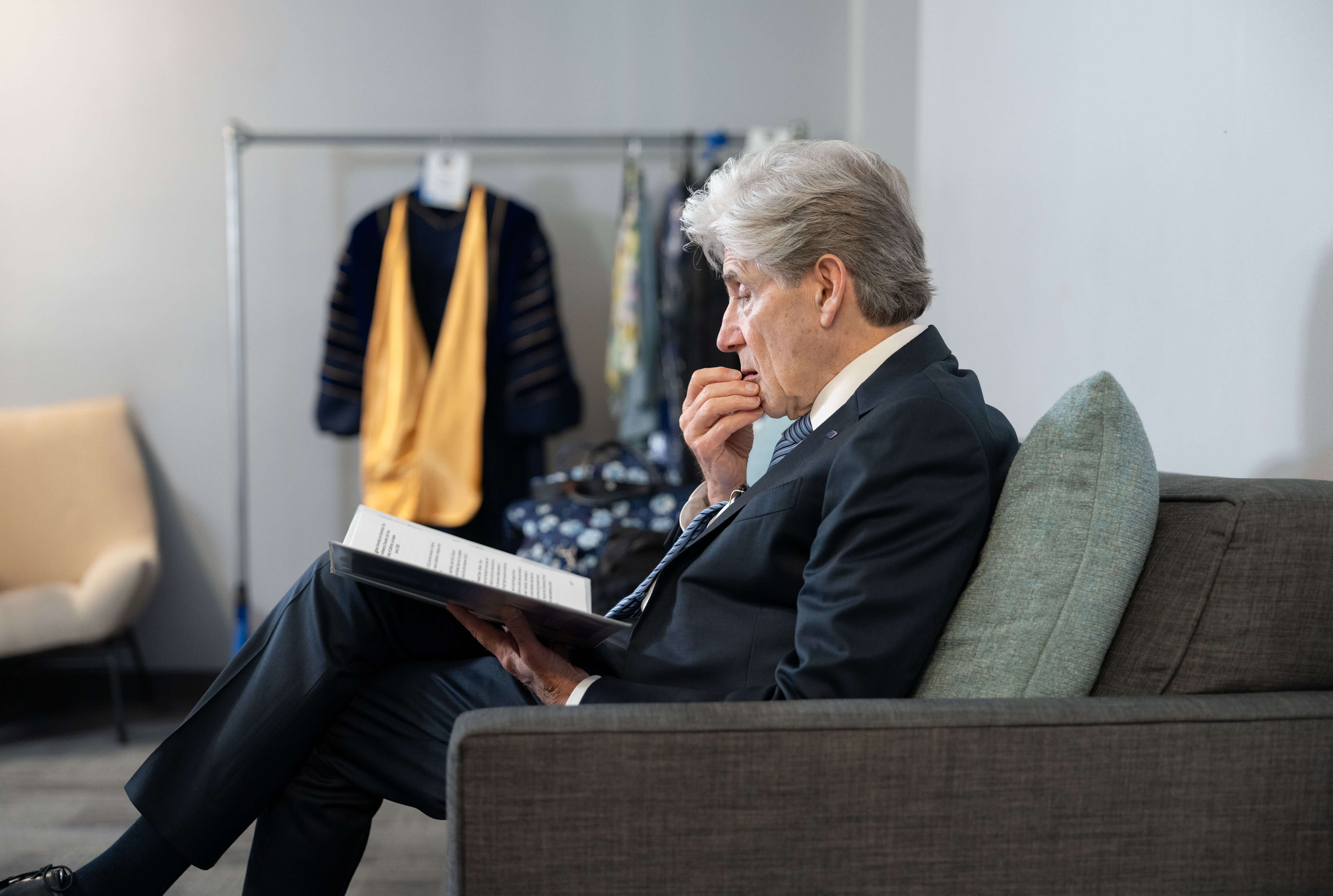 Chancellor Frenk in a suit and tie is seated on a couch reading from a book