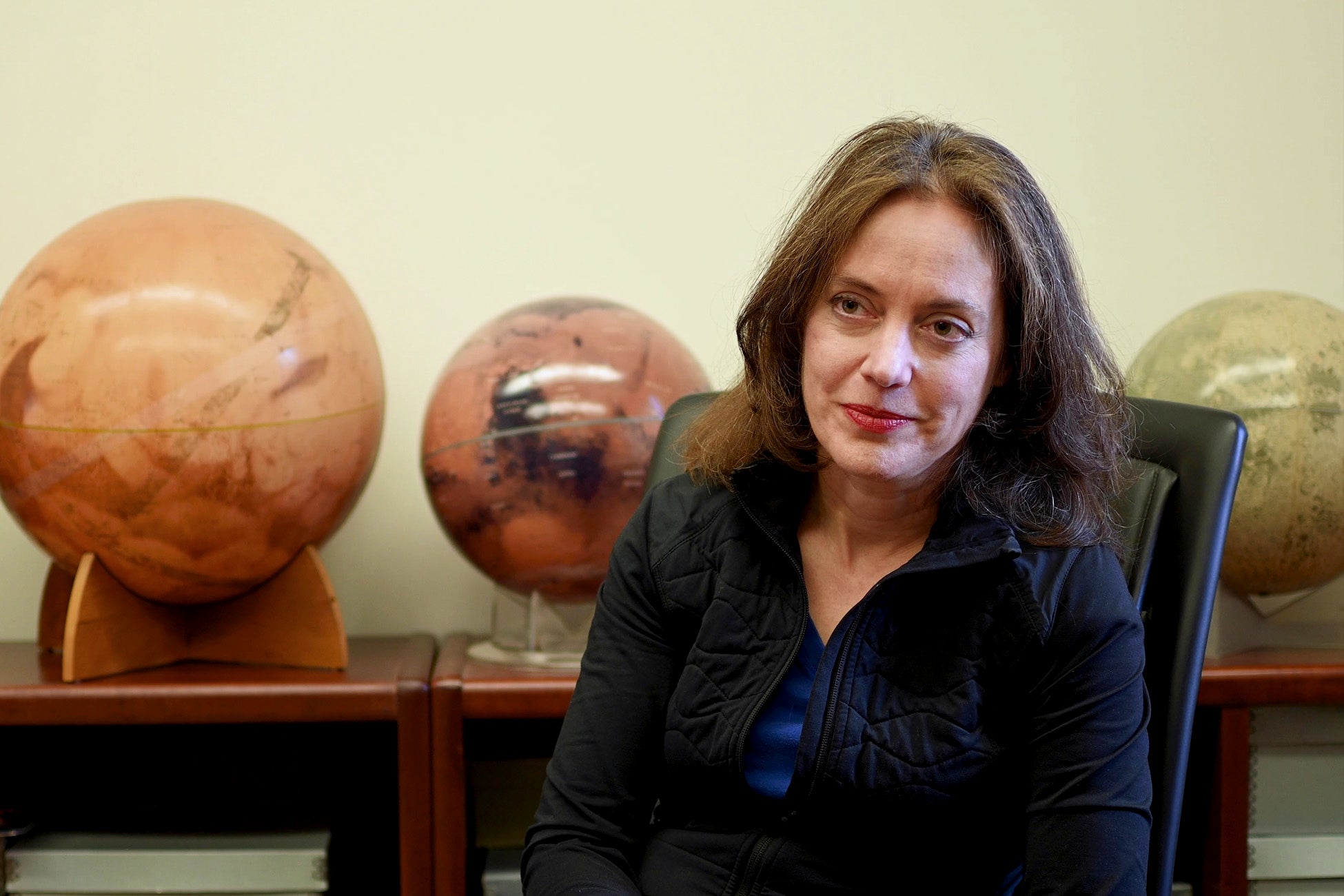 A woman sits in an office chair, looking slightly to the side, with three spherical Mars globes displayed on shelves behind her.