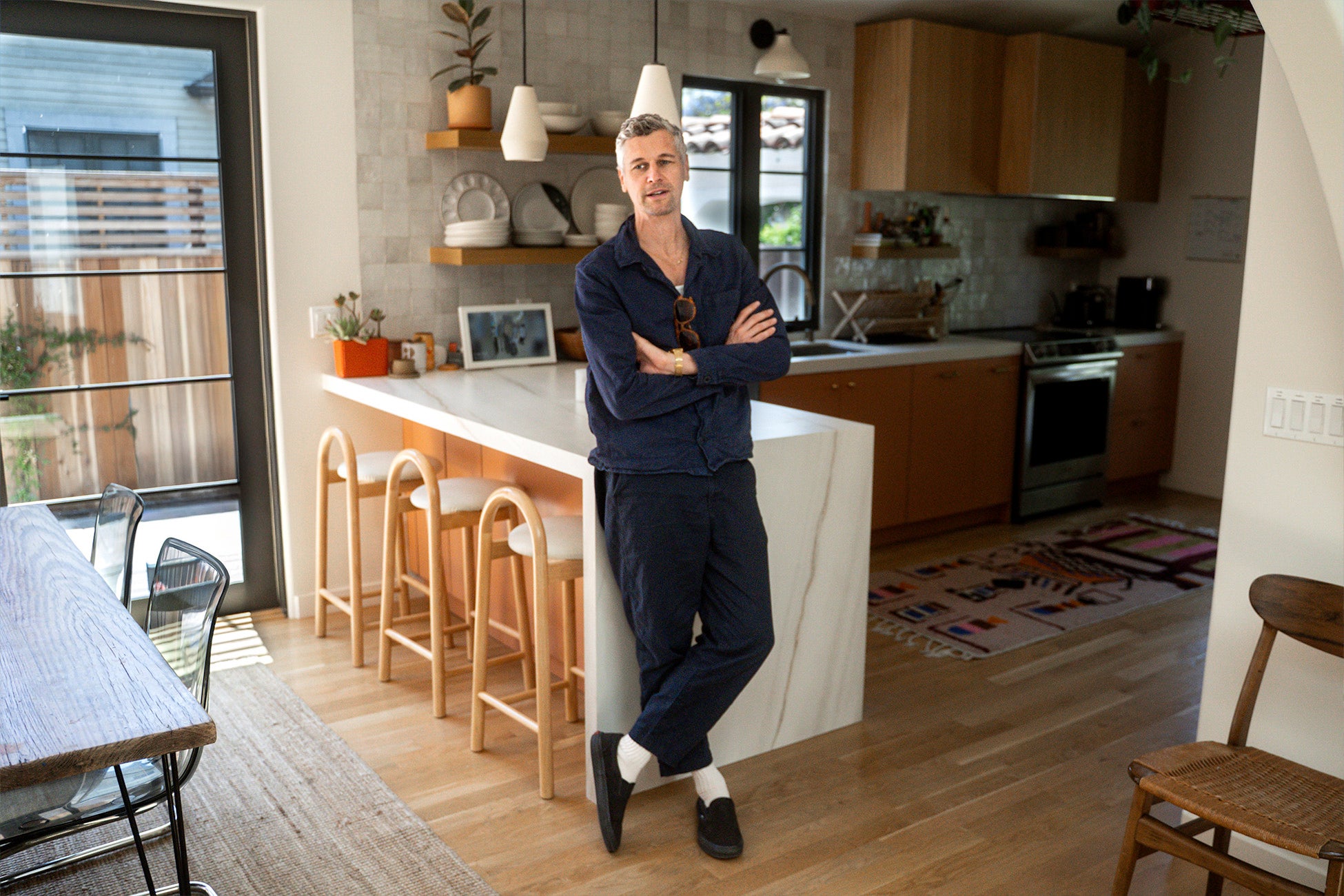 A man stands with his arms crossed, leaning against a white kitchen island in a bright, modern home. Pendant lights and bar stools are visible behind him.