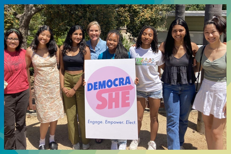 A group of women stand smiling gathered around a sign reading "Democrat She, engage, empower, elect".