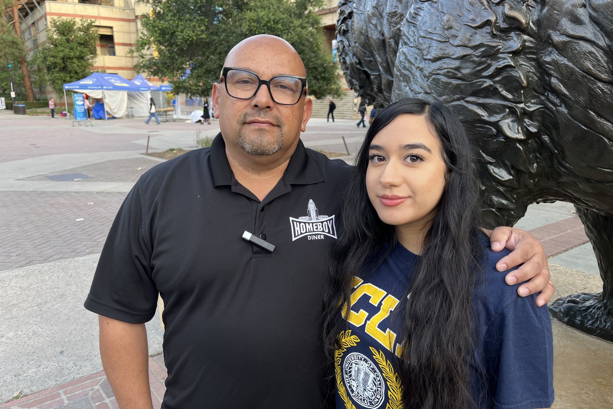 A man with glasses and black shirt stands with his arm wrapped around a young woman with long dark hair and blue UCLA shirt. Both stand beside the bruin bear statue with students and ackerman union building in the distance.
