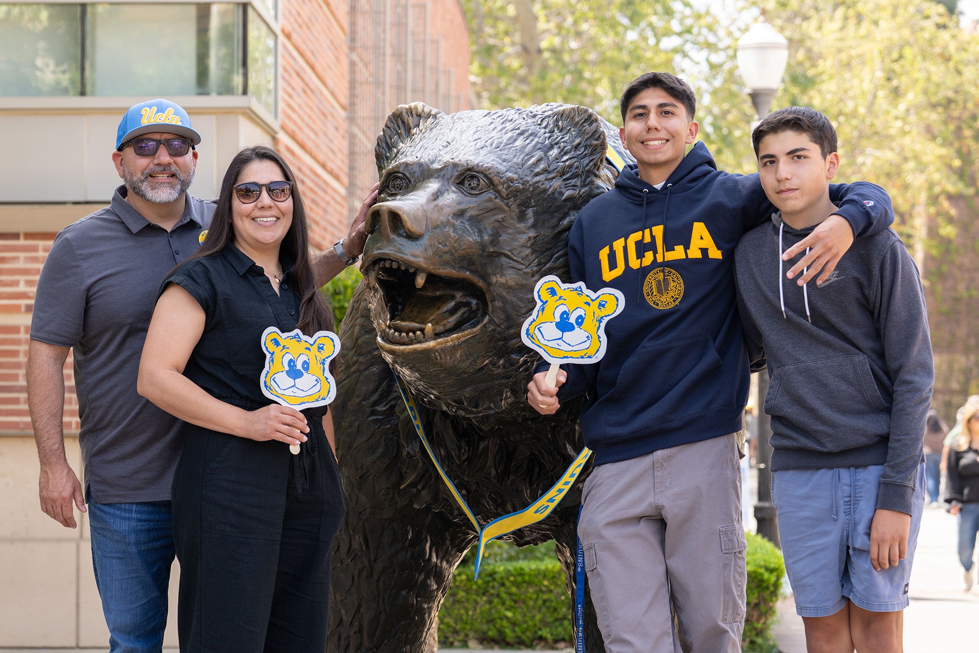A dad, mom and two sons, one doing a “4s up” hand gesture, pose in UCLA swag next to The Bruin statue.