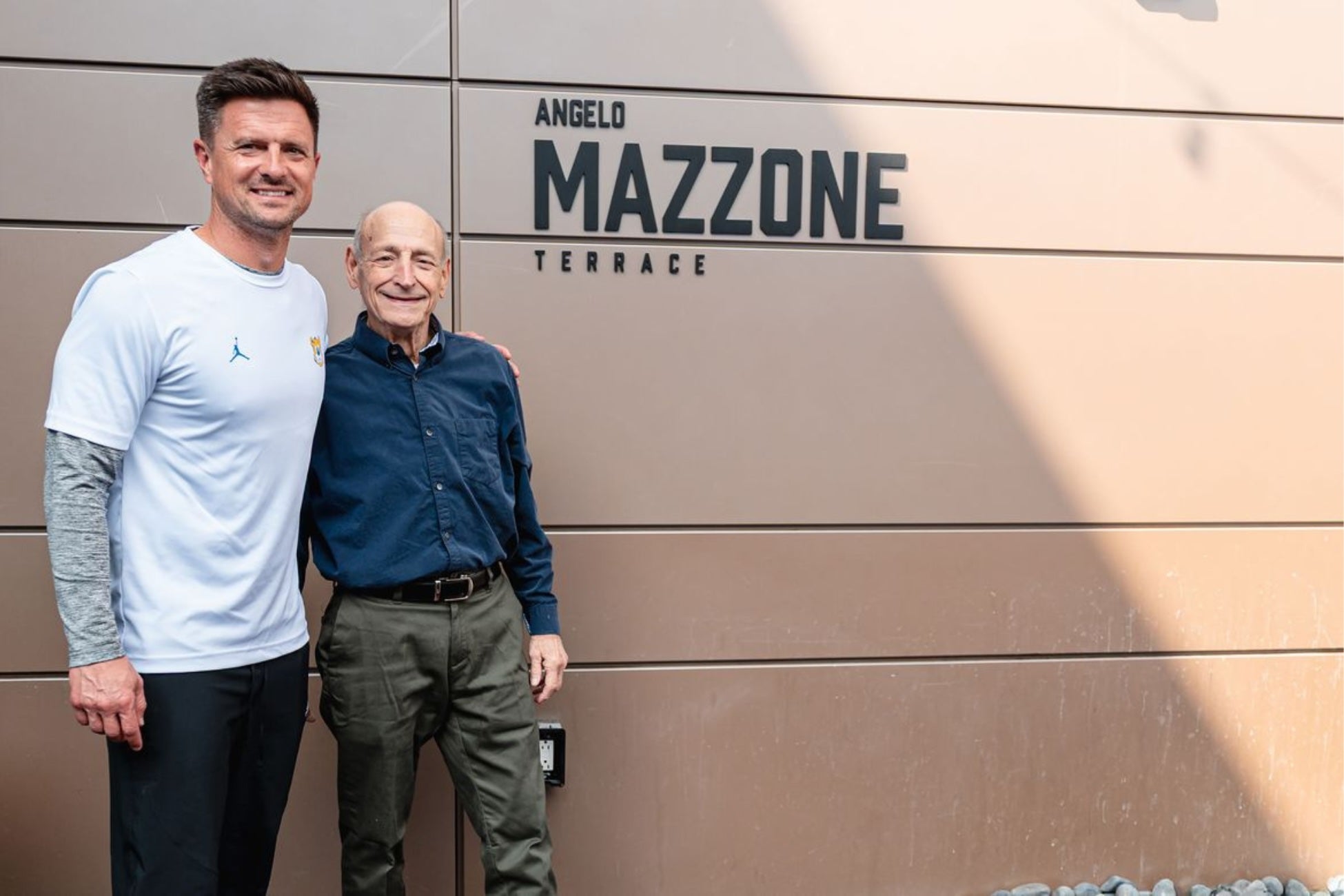 Two men stand in front of a tan building smiling with the words "Angelo Mazzone Terrace". One man is wearing a white shirt and black pants. The other man is wearing a blue button up shirt with dark pants.  