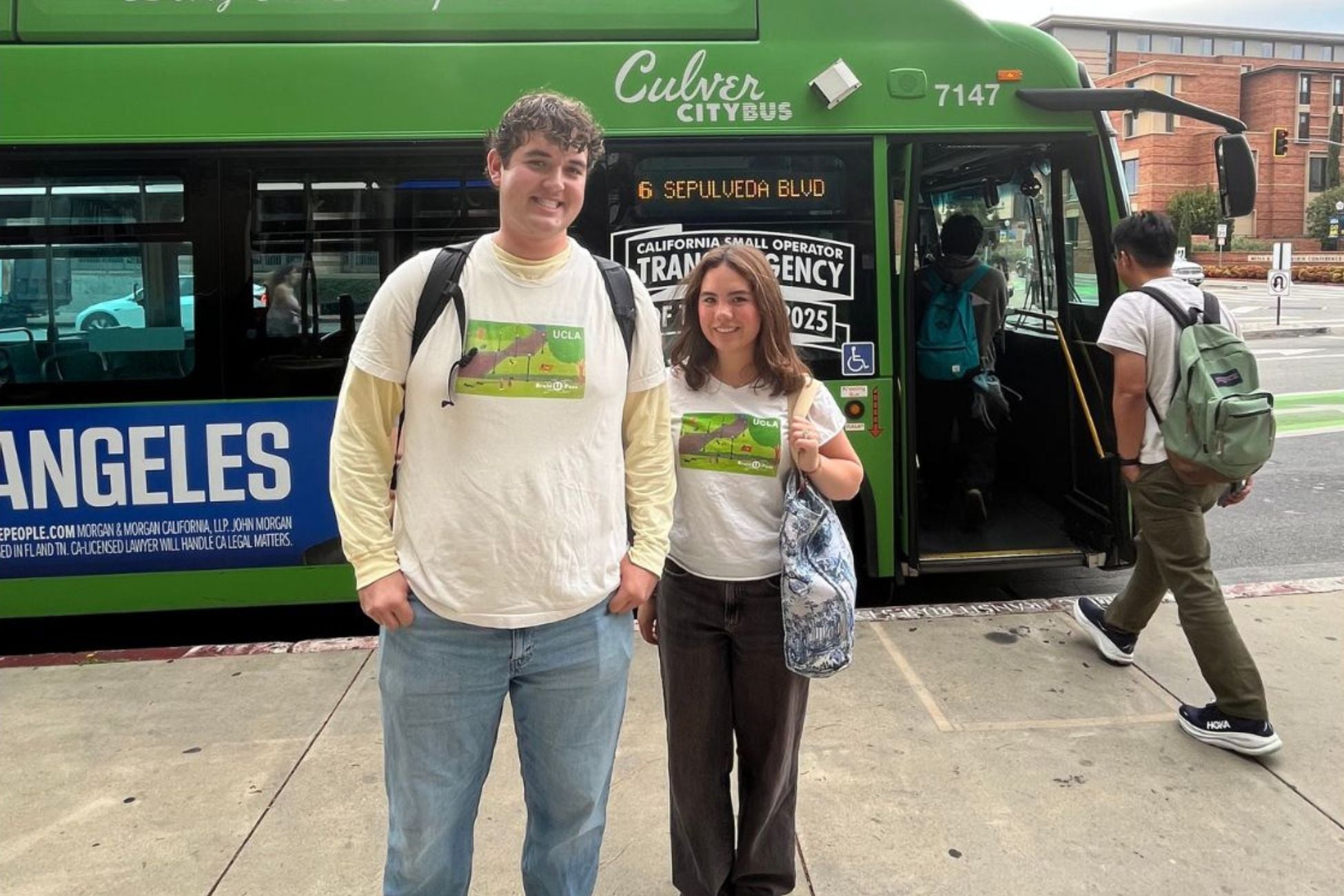 A man and woman stand together in front of a green bus, smiling and dressed casually.