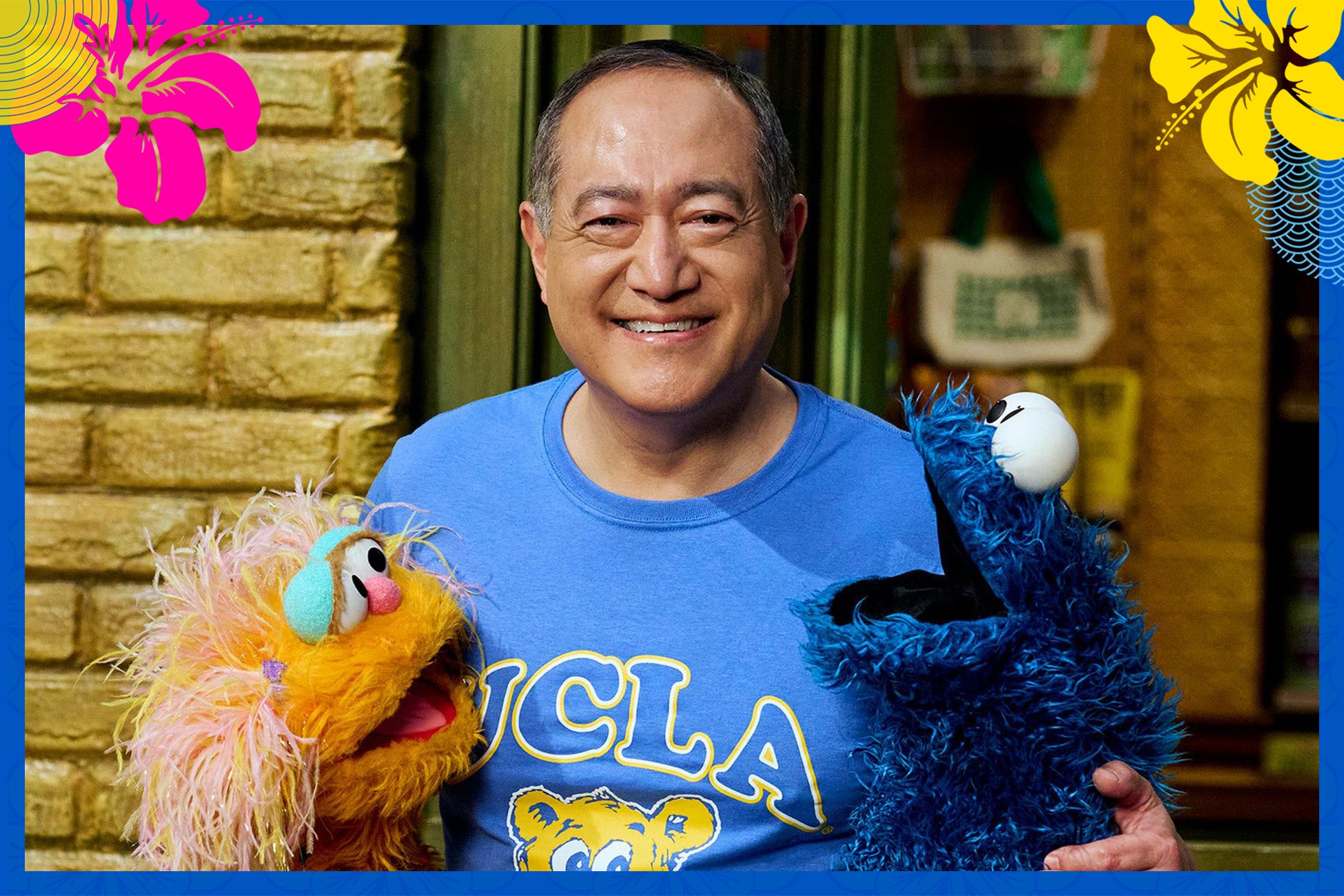 A man in a blue UCLA shirt holds two muppets, one blue and one yellow, smiling at the camera.
