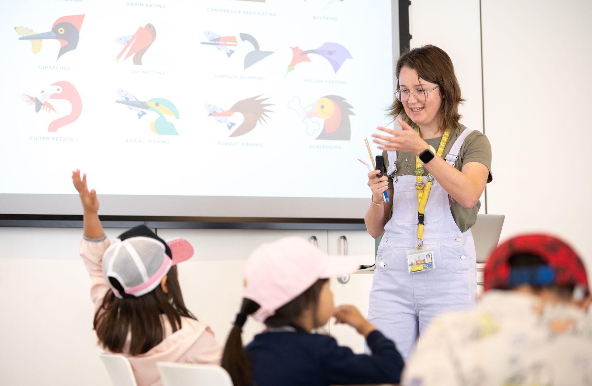 A young woman in overalls stands in front of a group of eager children in a classroom.