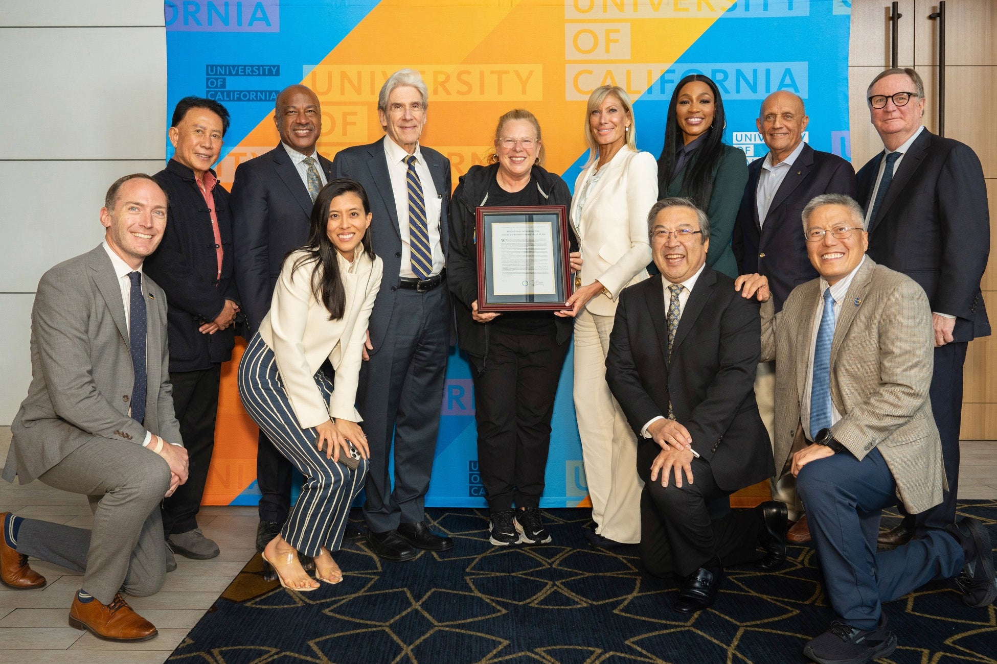 A group of eleven people poses in front of a University of California branded backdrop. A woman in the center holds a framed certificate.