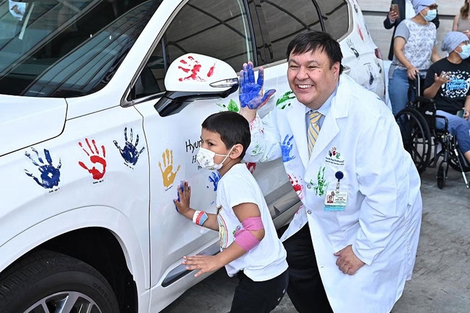 A man in a lab coat stands smiling with a hand covered in paint. A child stand beside him next to a car that is covered in color handprints. The child is pressing his hand into the car, leaving a color handprint.