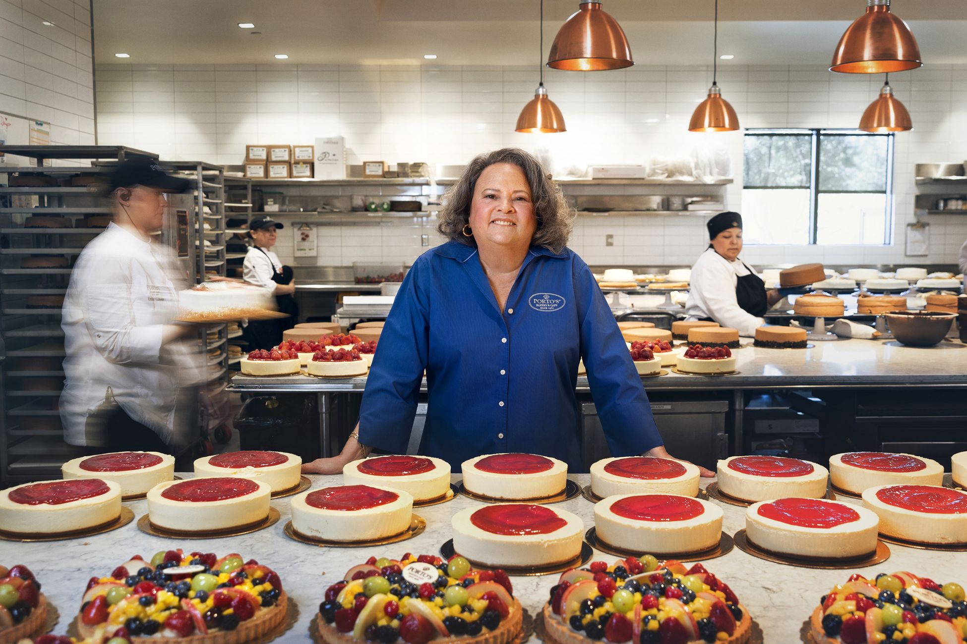 A woman stands smiling in front of a large display showcasing various cakes in a bakery. 