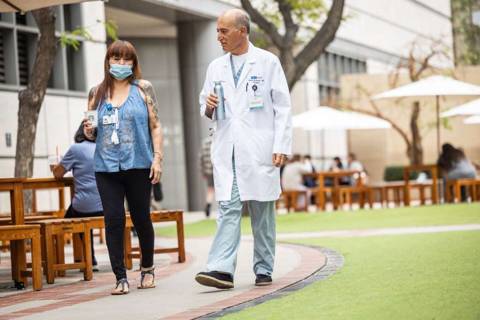 A man, wearing a white lab coat over scrubs, walks outdoors with woman, who is wearing a protective mask