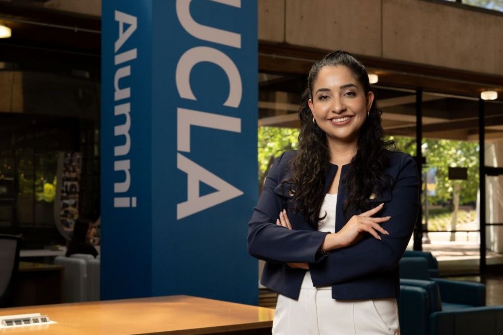 Cinthia Flores, UCLA Alumni Association President, standing with arms crossed beside a UCLA Alumni sign indoors.