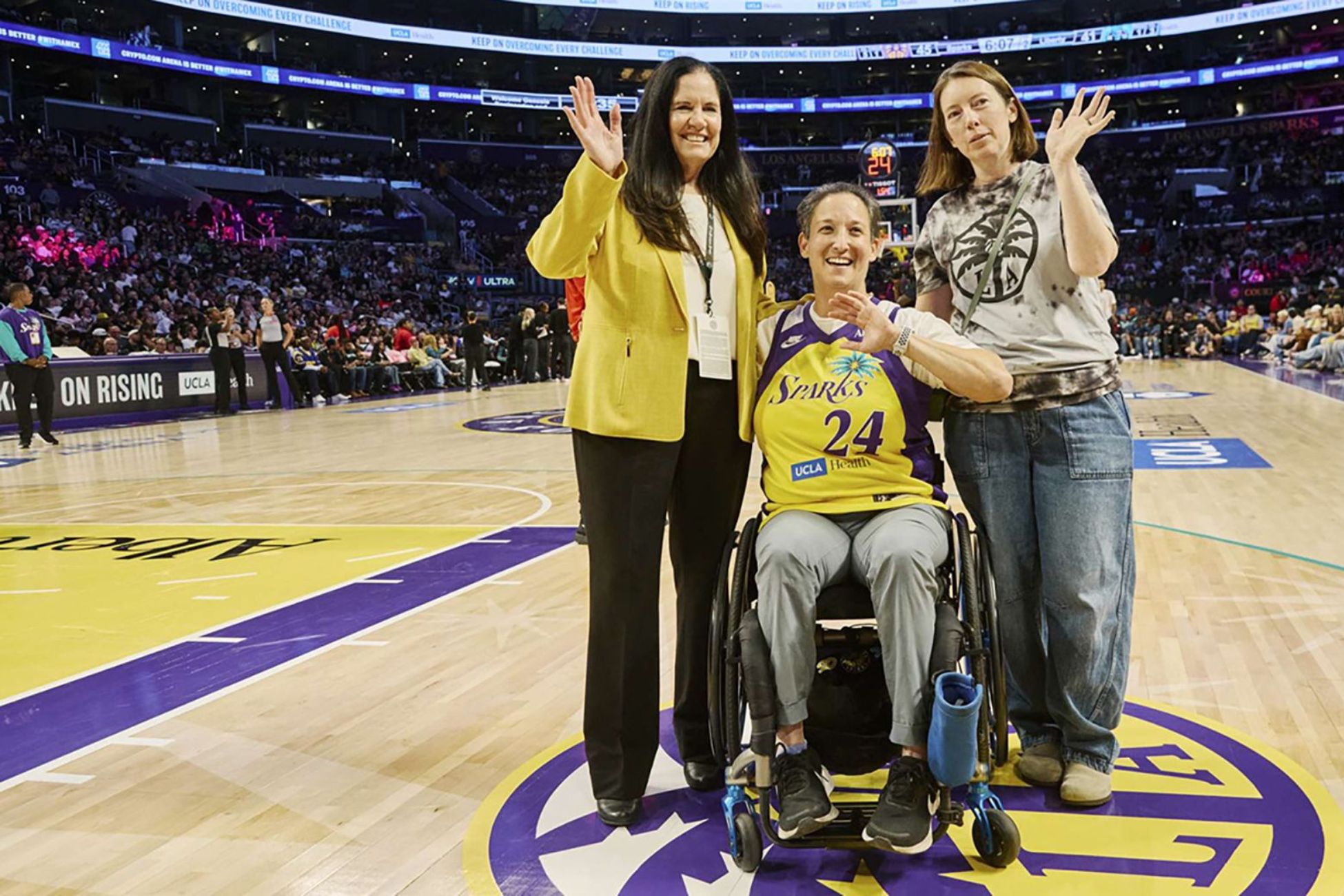 From left: Sharon Hame, in a yellow jacket and black pants, Laura Goodkind, seated in a wheelchair and wearing a yellow and purple Sparks jersey, and Jennie Coyne, in a graphic T-shirt, pose courtside at a basketball arena.