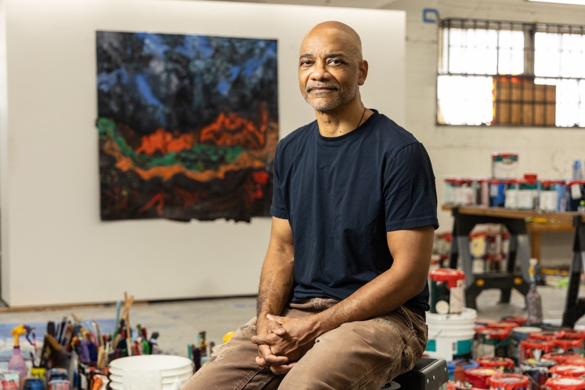 Rodney McMillian sits in his art studio, surrounded by paint cans, brushes and other supplies, with a large abstract painting hanging behind him.