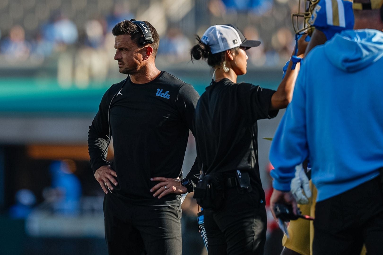 Coach Bob Chesney stands focused on the field wearing a UCLA shirt and headset during a game.