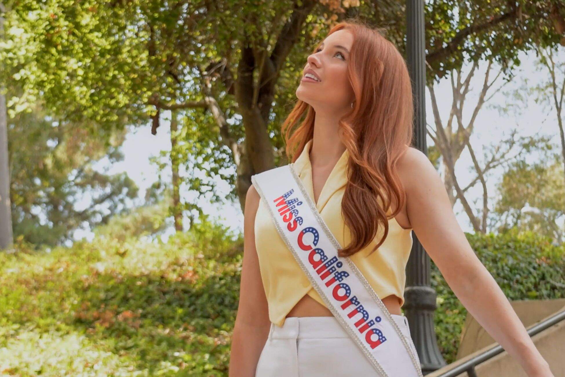 Sharlize True Trujillo wearing a Miss California sash looks up thoughtfully while standing outdoors near trees and stairs.