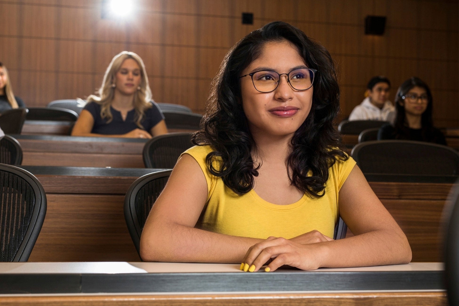 Students observe proceedings in a wood-paneled courtroom, with a woman in a yellow top and glasses seated in the foreground