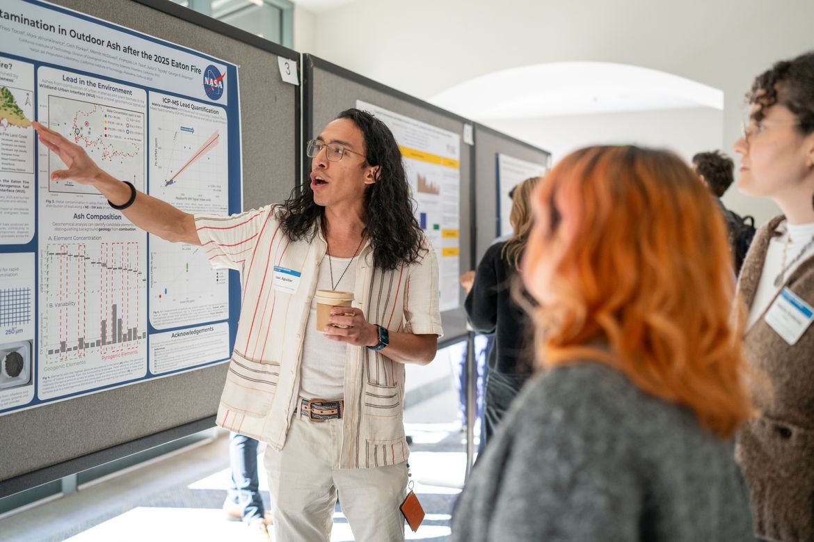 Isaac Aguilar, in white shirt and pants, points at a poster while speaking