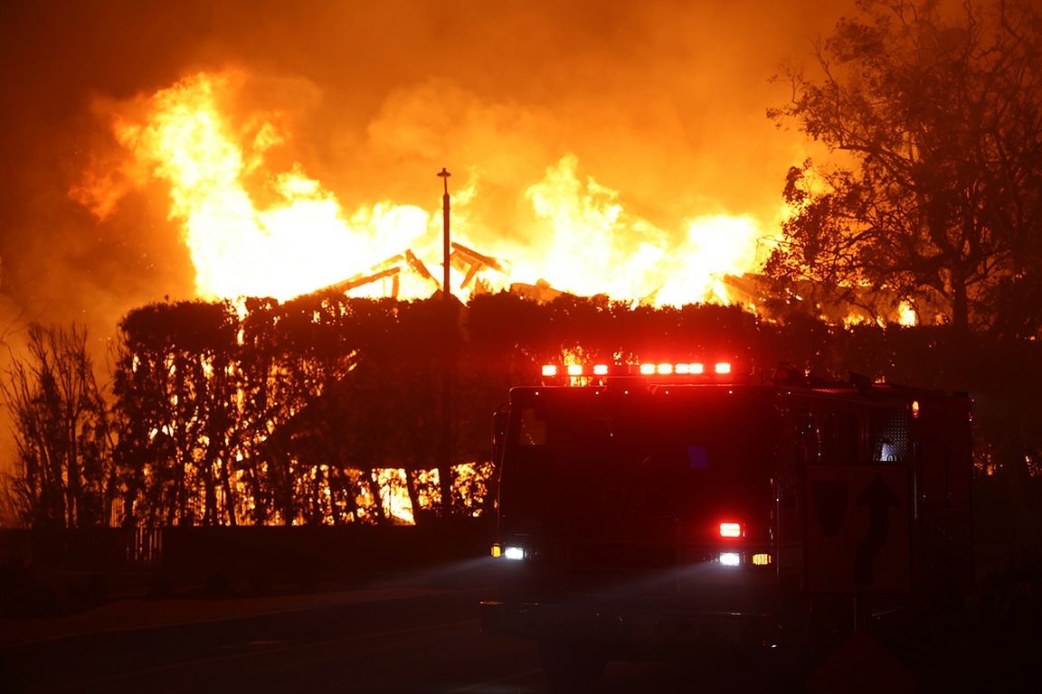 Fire truck with lights flashing parked before a large structure fire at night, flames and smoke filling the sky.