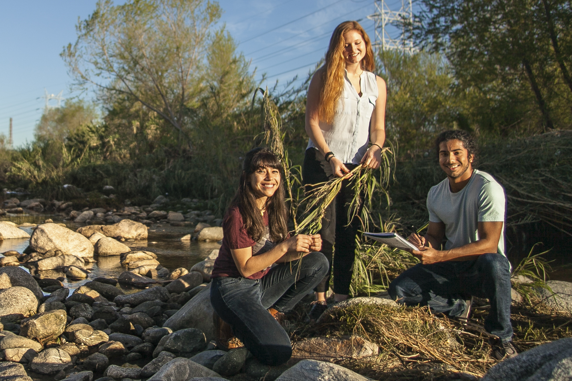 A female with long hair stands upright holding broken branches in her hands while another female and a male crouch down collecting additional branches and recording their work on paper as they clean up the Los Angeles River