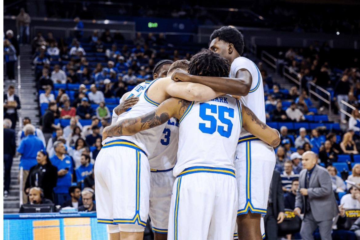 UCLA men’s basketball players in white and blue uniforms huddle together on the court during a game, arms around each other in a show of teamwork and focus.