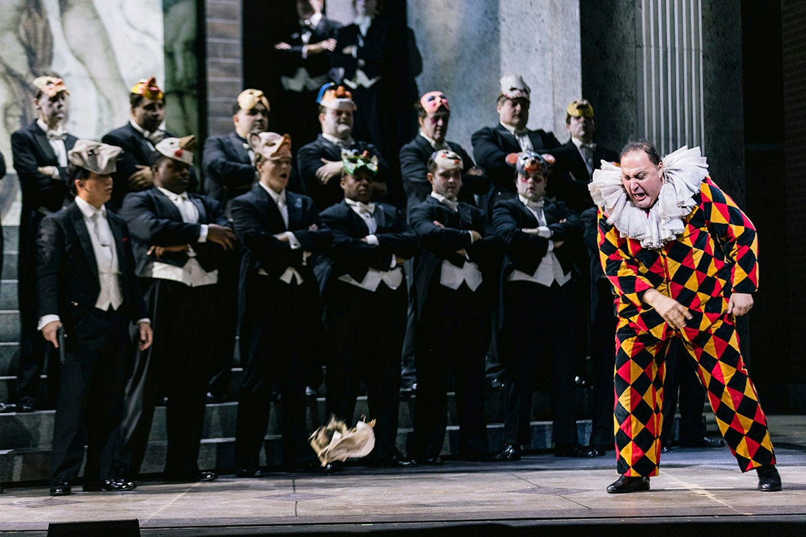The jester Rigoletto performs in front of a crowd of masked men, judging with their arms crossed.