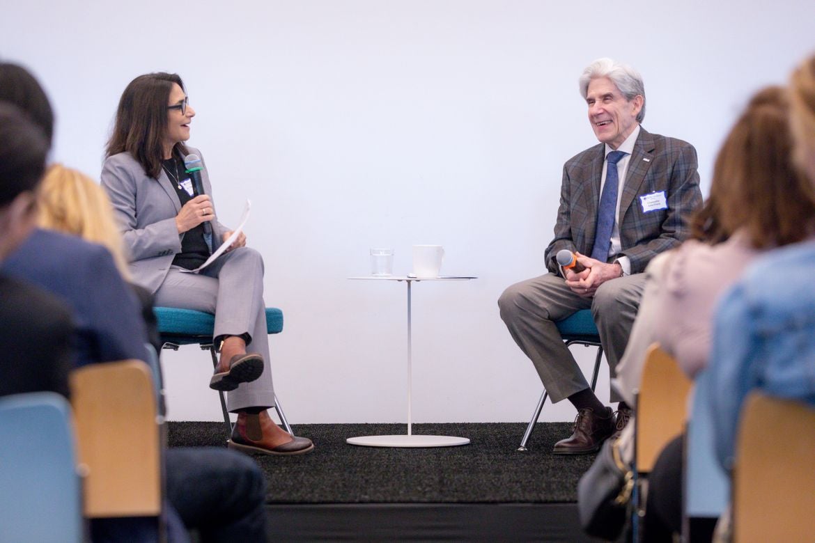 Tina Christie, left, in gray suit, and Julio Frenk, in brown suit and blue tie, hold microphones while seated in front of audience