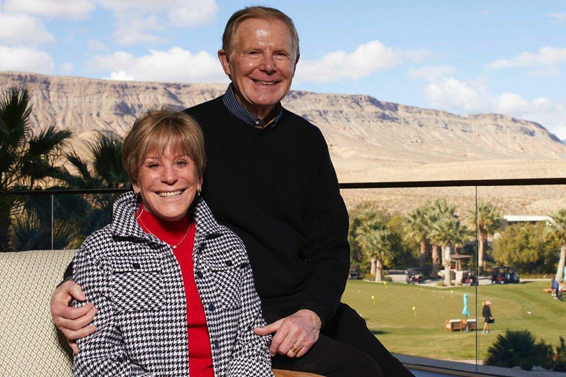 Heidi Canarelli, left, in red, black and white, next to Larry Canarelli, in black sweater, with golf course, palm trees and mountains in background
