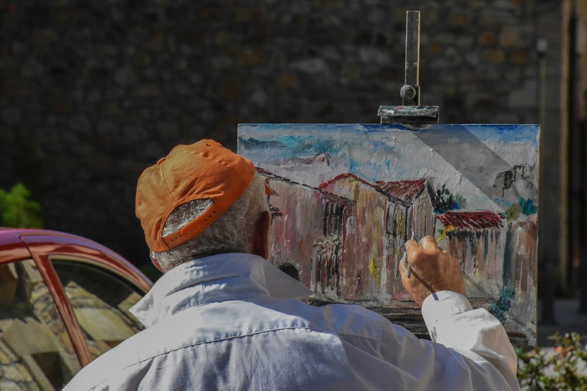Older man in red hat paints a picture of a building on a canvas sitting on an easel