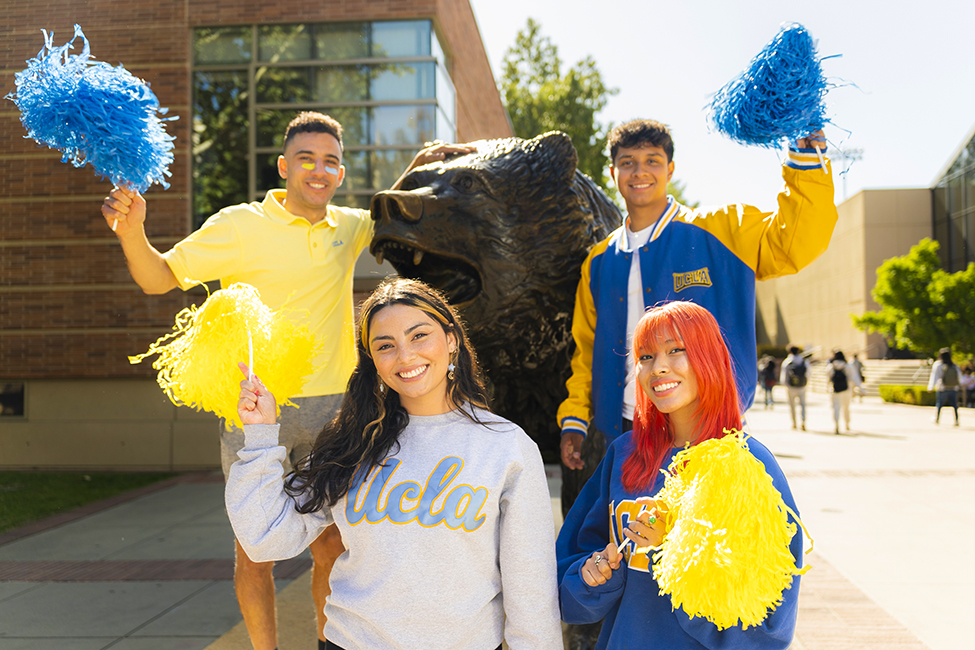 Four students holding blue and yellow pompoms pose with the Bruin Bear statue