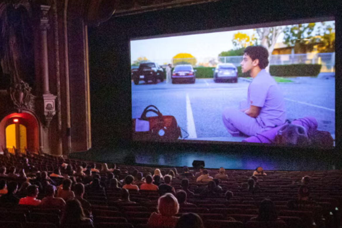 Audience in the ornate LA theater watching a film. On screen is a man sitting in a parking lot with a luggage and yogamat next to him.
