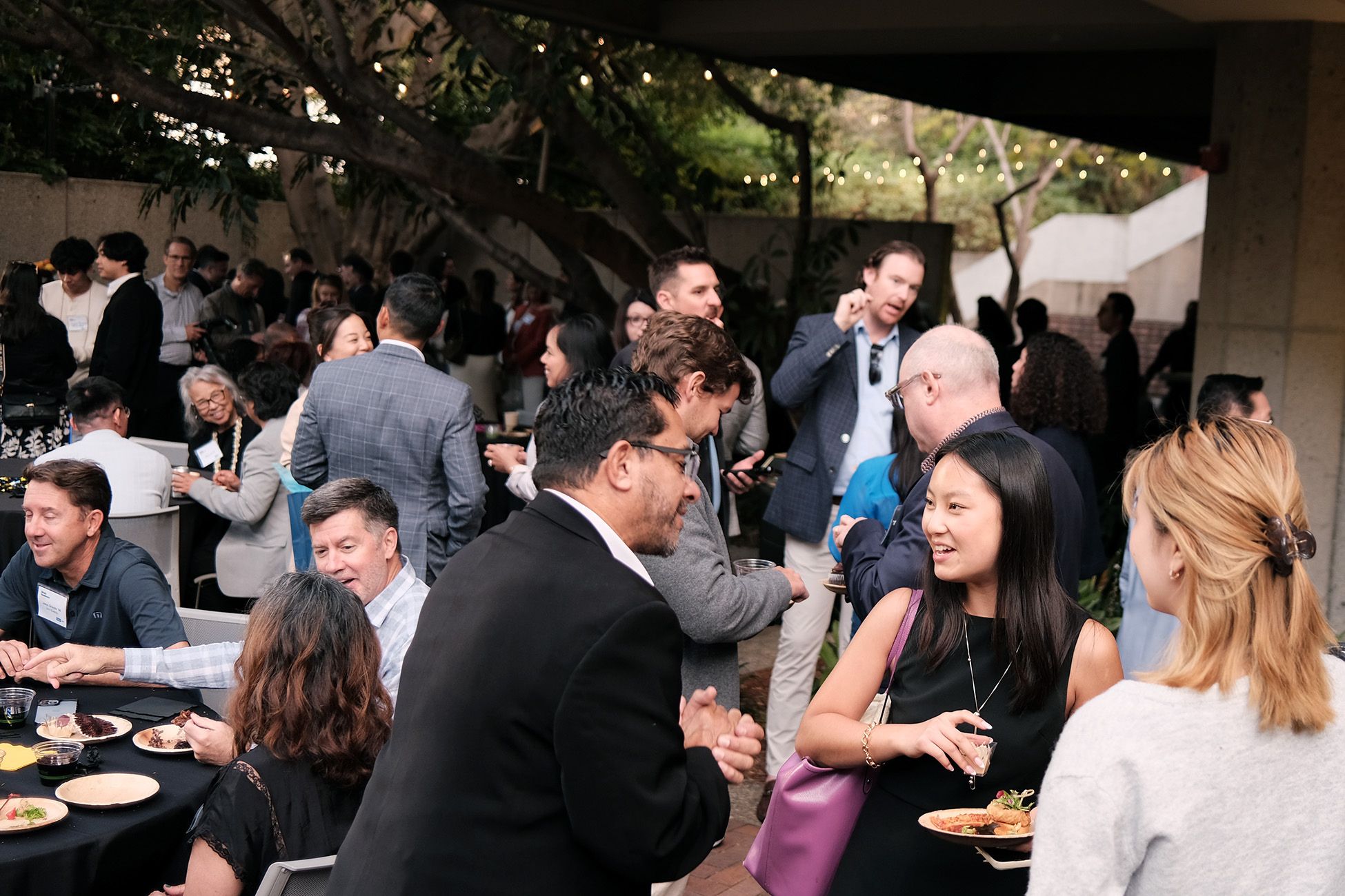A large group of people mingle and chat at an outdoor networking event, surrounded by string lights and shaded by trees. Some attendees are seated at tables enjoying food, while others stand and engage in conversation, holding plates and drinks.